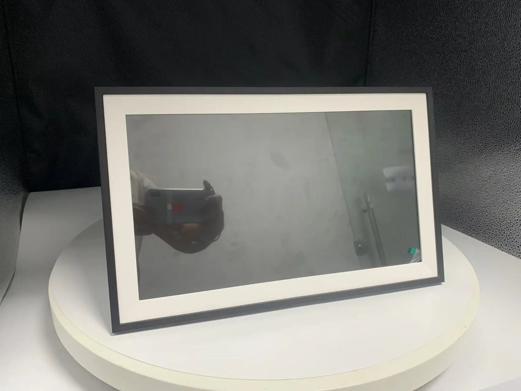 White 15.6 inch smart digital photo frame on a table showing a family photo, with mobile app upload icon.