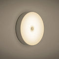 Small round white LED night light with motion sensor and USB charging cable on a table.