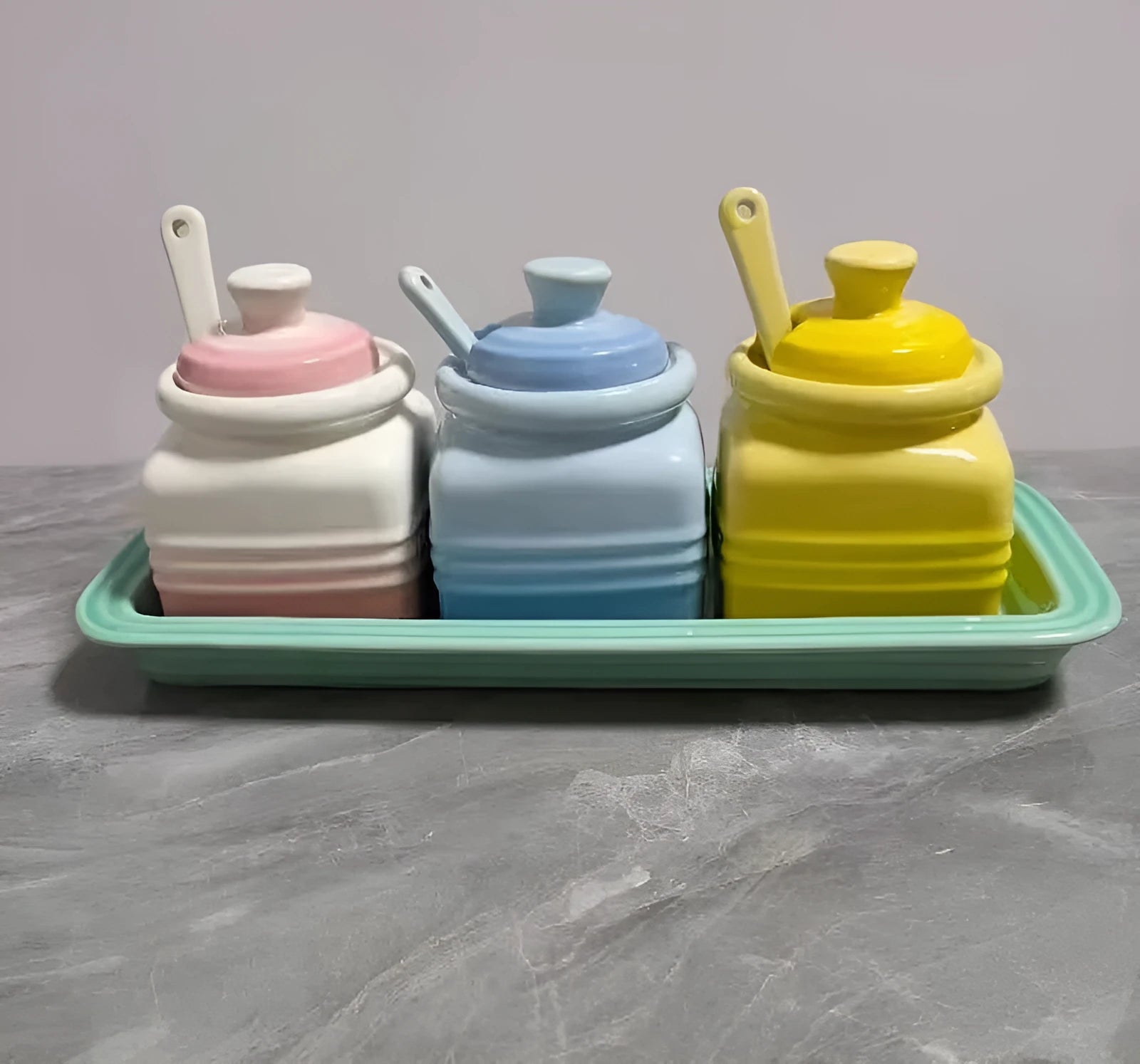 Three cool-coloured ceramic seasoning pots with a matching spoon on a kitchen counter