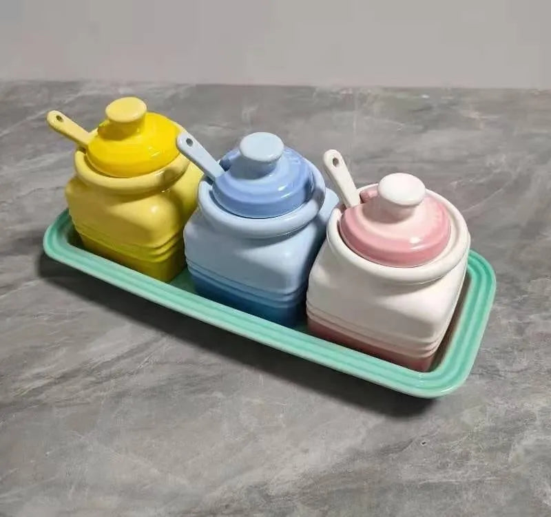 Three cool-coloured ceramic seasoning pots with a matching spoon on a kitchen counter