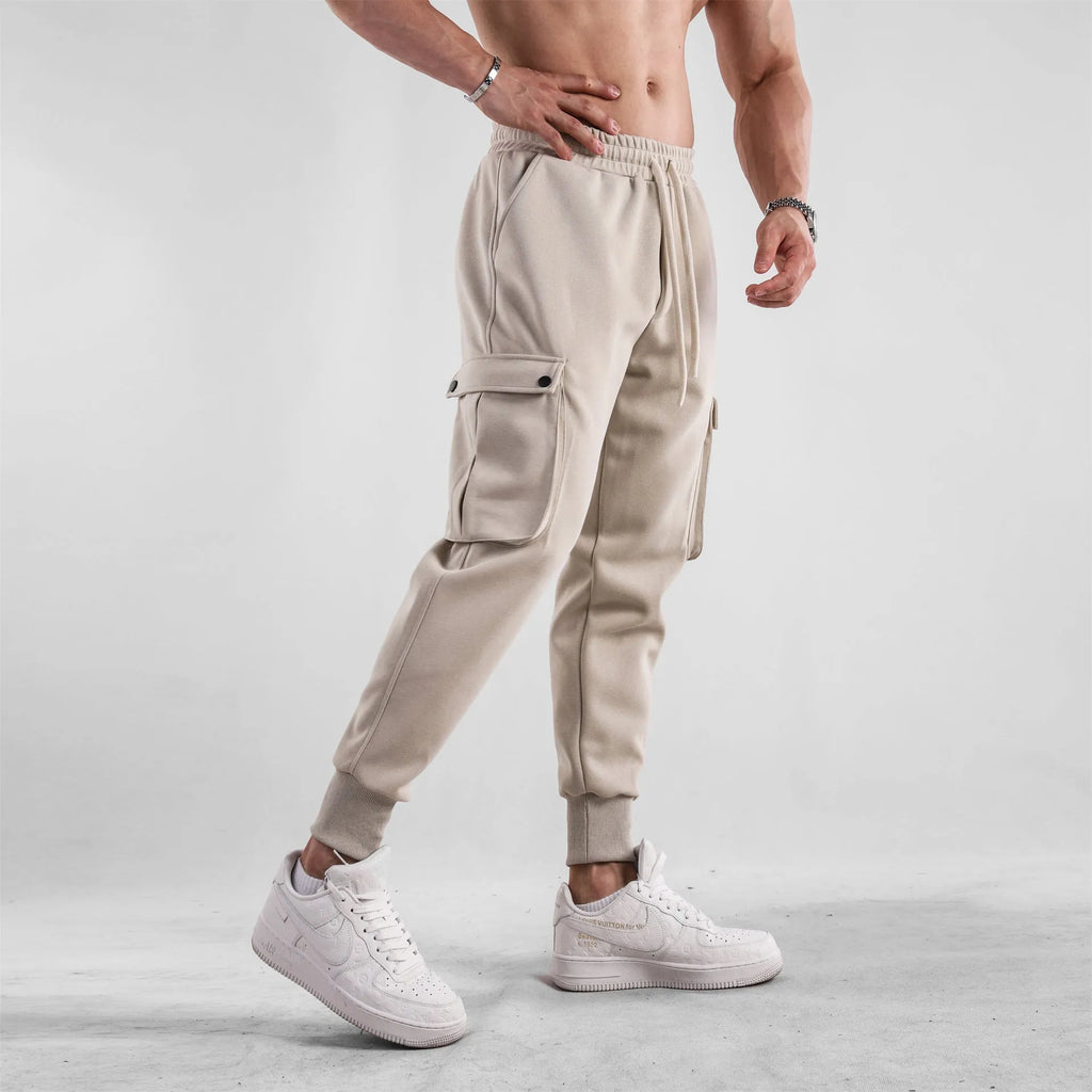 Men's loose-fit beige cotton sports trousers with ankle ties and side pockets laid flat.