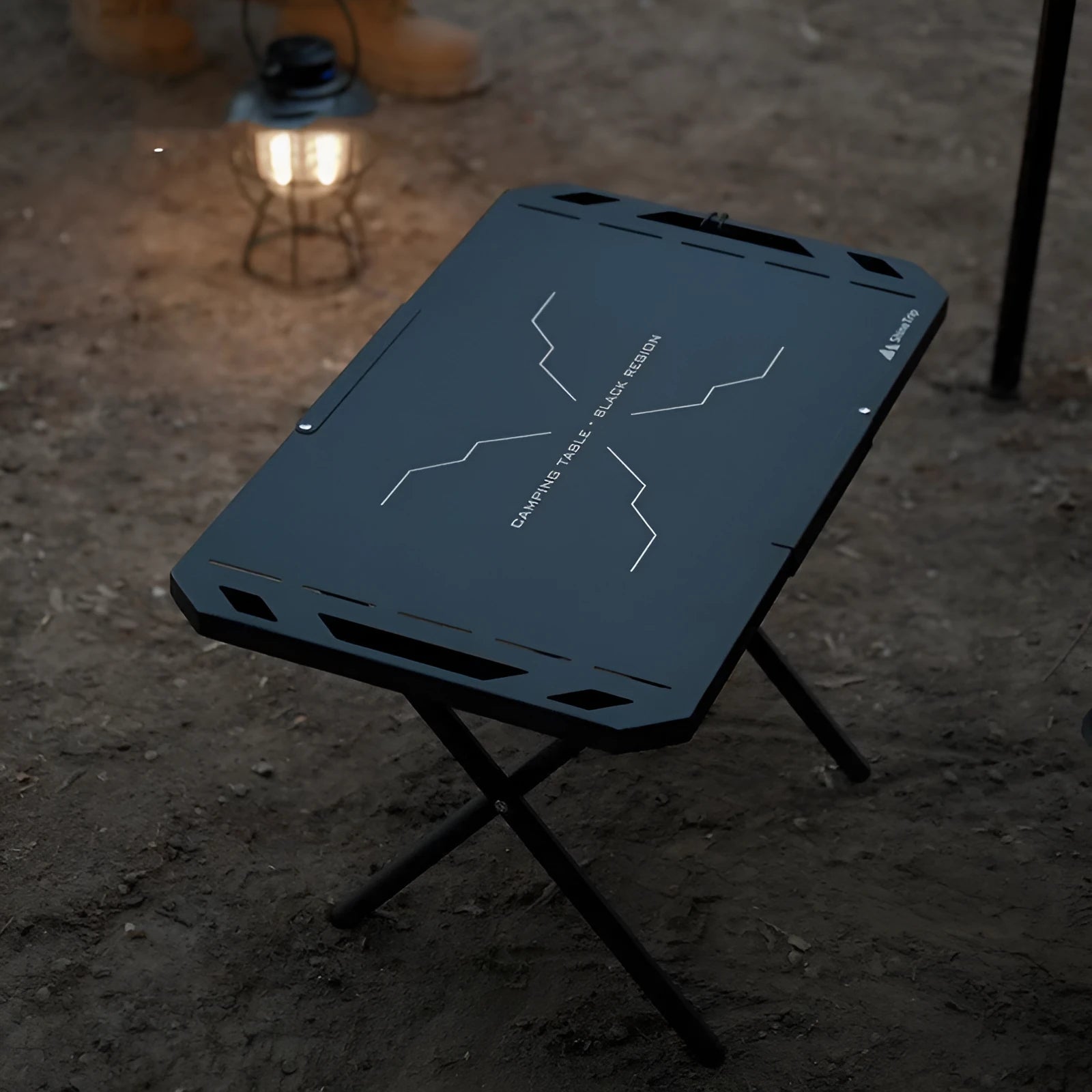 Black folding portable steel camping table on grass with storage bag beside it, 600x350x375mm dimensions visible