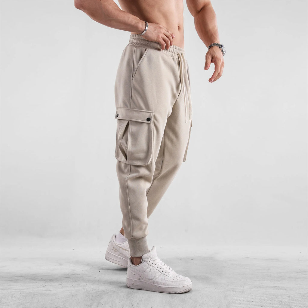 Men's loose-fit beige cotton sports trousers with ankle ties and side pockets laid flat.