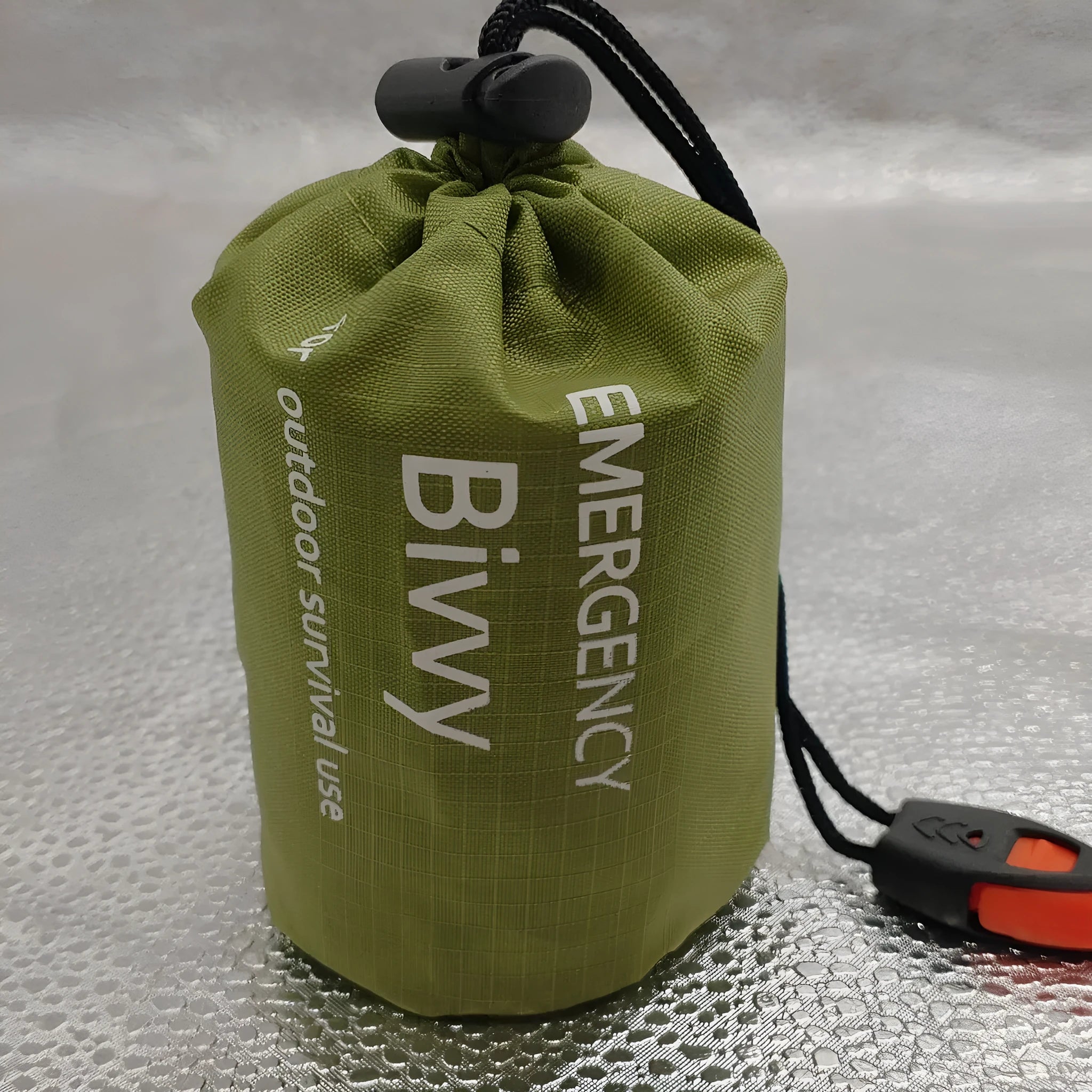 Orange aluminium foil emergency sleeping bag with drawstring bag and whistle on ground