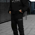 Man wearing a grey hooded sweater tracksuit with letter print on the chest, featuring matching trousers.