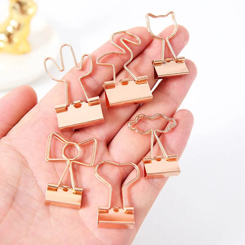 Gold metal decorative paper clips in shapes of a flamingo, a cat, a cactus, a bow, a love symbol, and clouds.