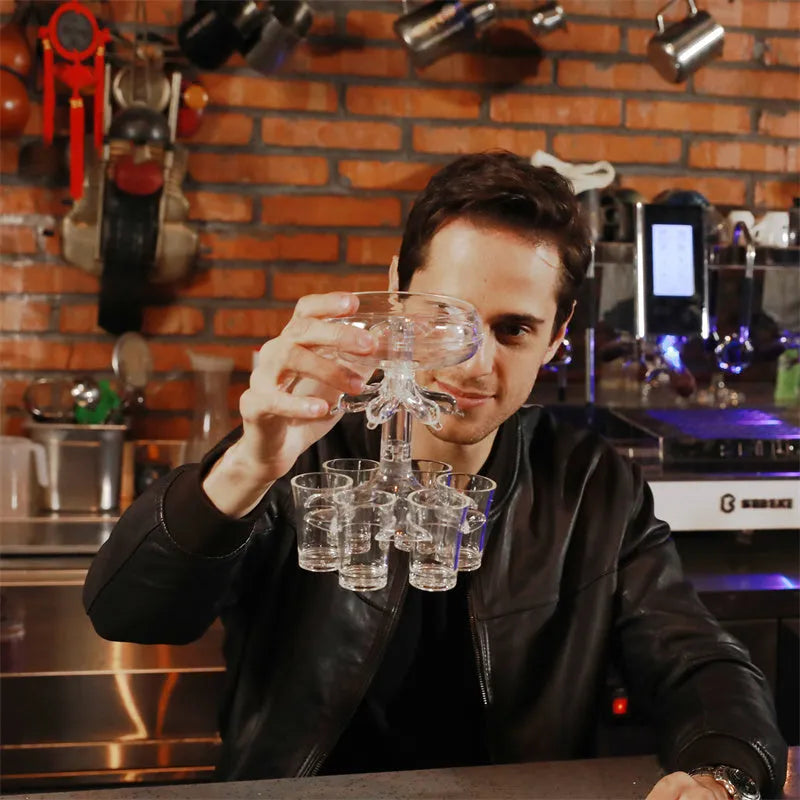 Transparent six-cup shot glass dispenser rack with gold-trimmed cups on a table.