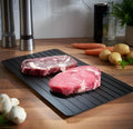 Black aluminium quick defrosting tray for thawing meat and frozen food on a kitchen counter.