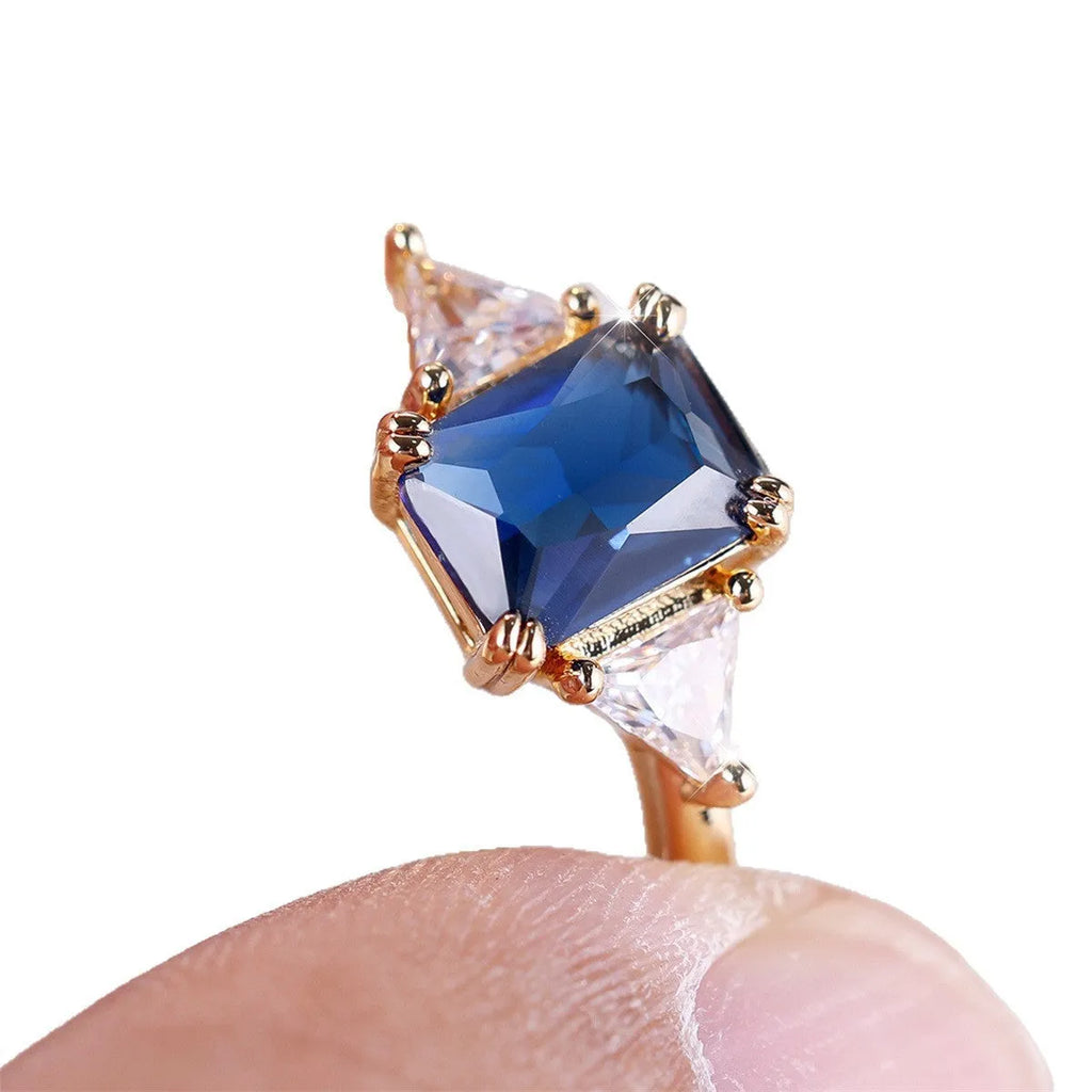 A rectangular gold sapphire blue plated copper ring for women on a plain background.