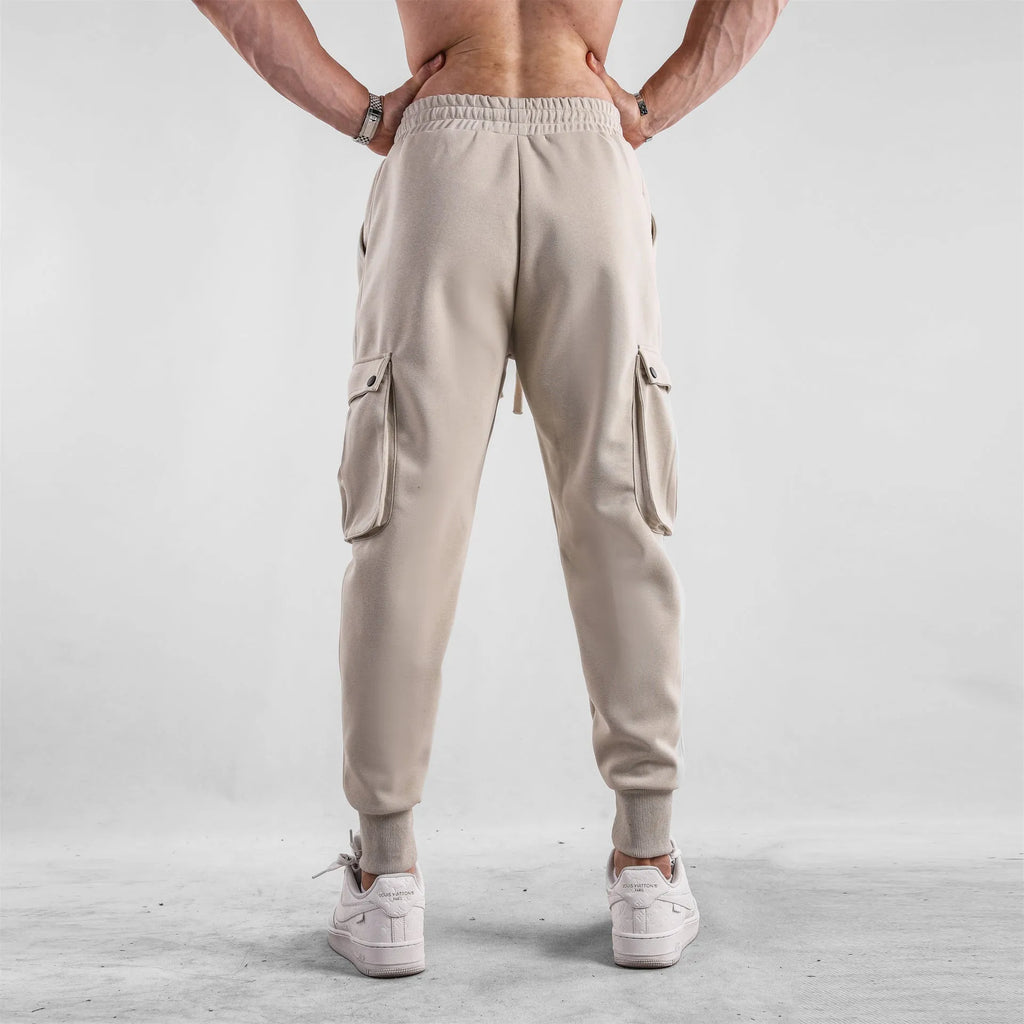 Men's loose-fit beige cotton sports trousers with ankle ties and side pockets laid flat.