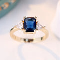 A rectangular gold sapphire blue plated copper ring for women on a plain background.