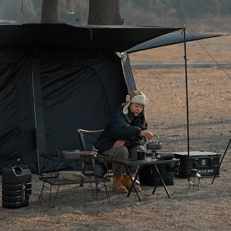 Black folding portable steel camping table on grass with storage bag beside it, 600x350x375mm dimensions visible