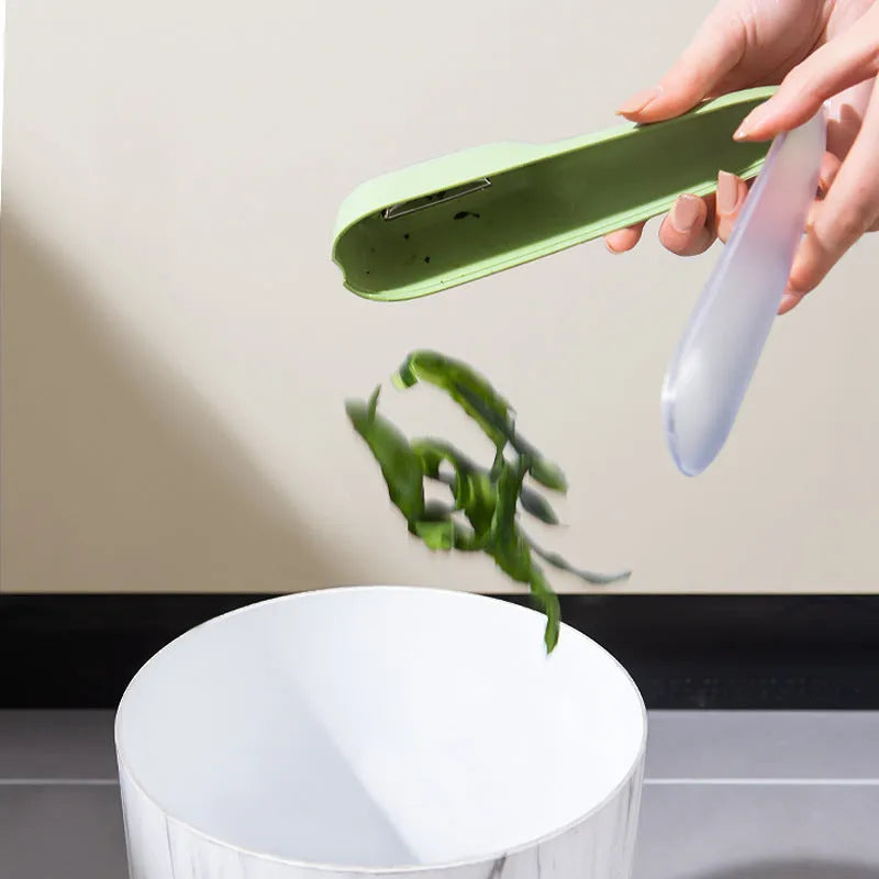 Dark green kitchen peeler with a storage tube, stainless steel blade, and non-slip plastic handle on a worktop.