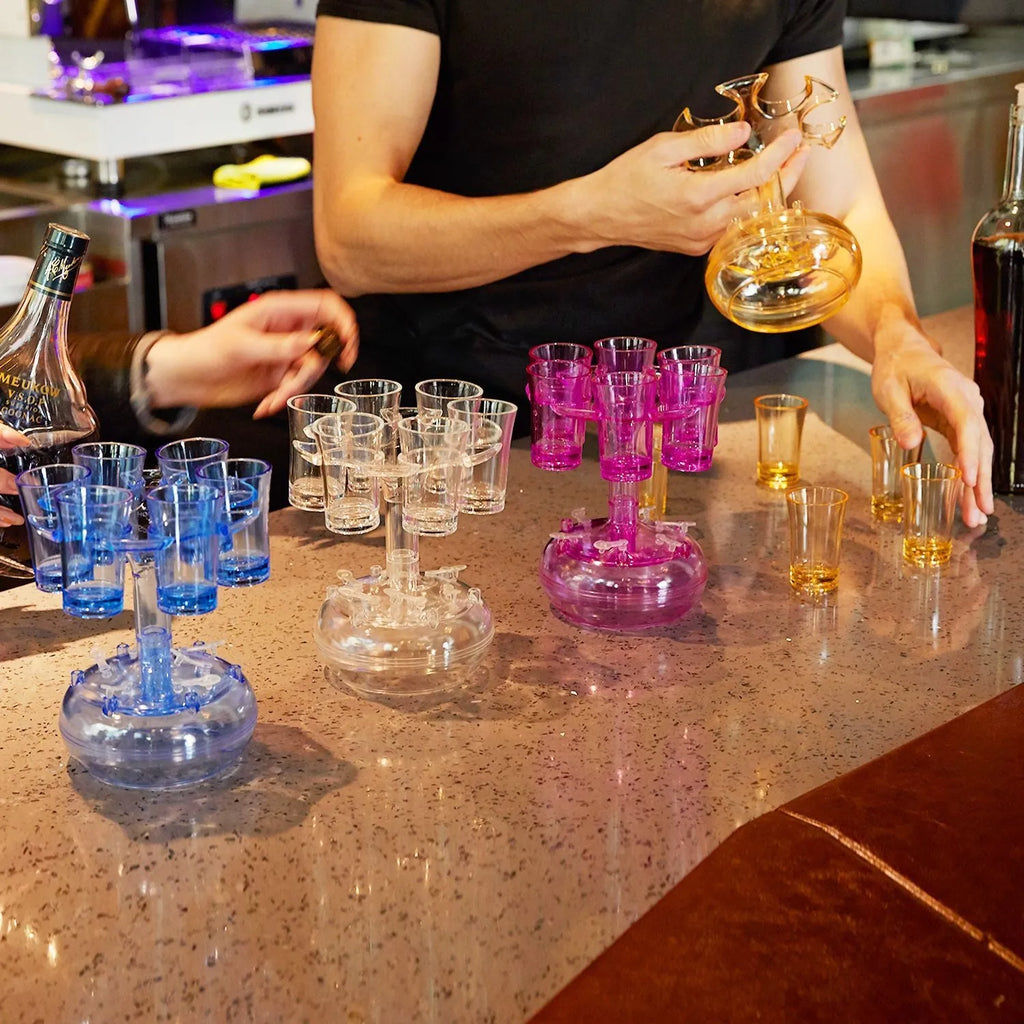 Transparent six-cup shot glass dispenser rack with gold-trimmed cups on a table.